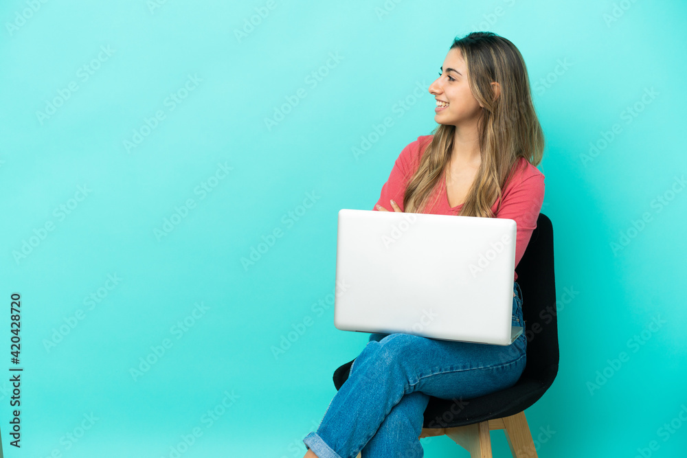 Young caucasian woman sitting on a chair with her pc isolated on blue background with arms crossed and happy Young caucasian woman sitting on a chair with her pc isolated on blue background with arms crossed and happy