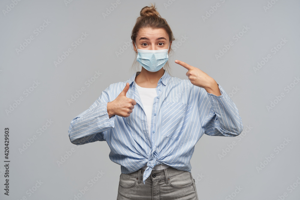 Cute girl with blond hair gathered in bun. Wearing striped knotted shirt and protective face mask. Pointing at the mask and showing thumb up sign. Watching at the camera, isolated over grey background Cute girl with blond hair gathered in bun. Wearing striped knotted shirt and protective face mask. Pointing at the mask and showing thumb up sign. Watching at the camera, isolated over grey background