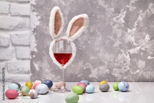 Glass of rose or red wine with bunny ears and Easter decorations, colorful eggs on white table, on bright background. Copy space for text.