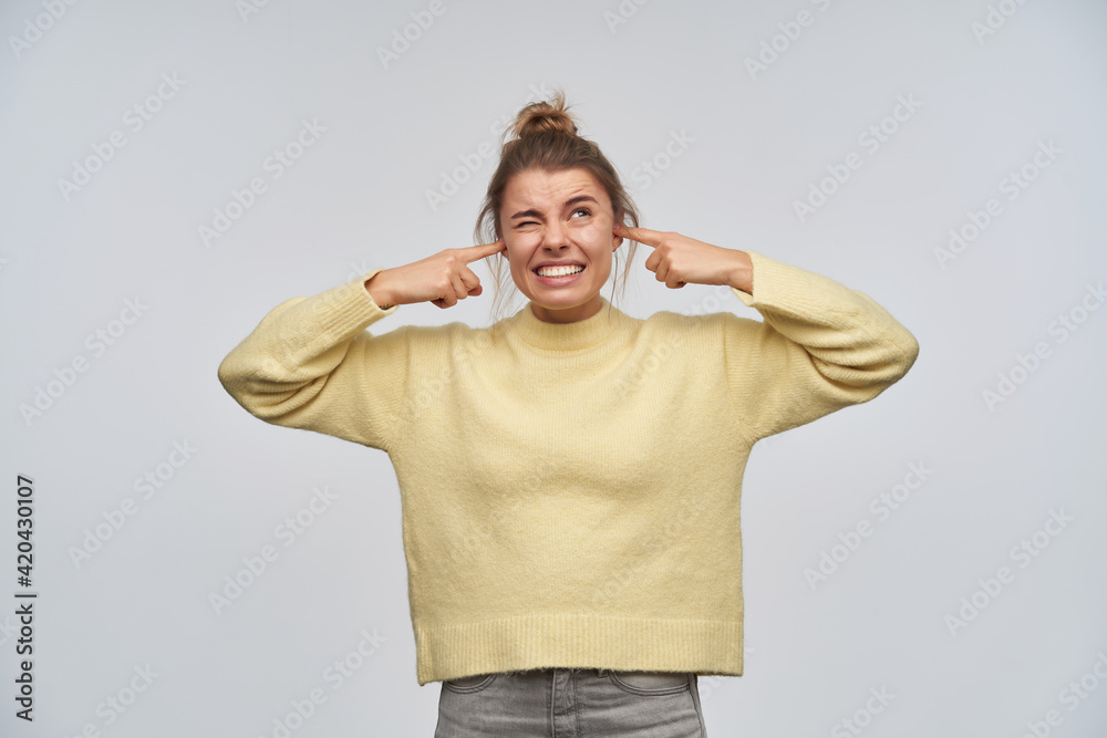 Annoyed looking woman, unhappy girl with blond hair gathered in bun. Wearing yellow sweater. Close her ears with fingers, it's too laud. Watching up at copy space, isolated over white background Annoyed looking woman, unhappy girl with blond hair gathered in bun. Wearing yellow sweater. Close her ears with fingers, it's too laud. Watching up at copy space, isolated over white background