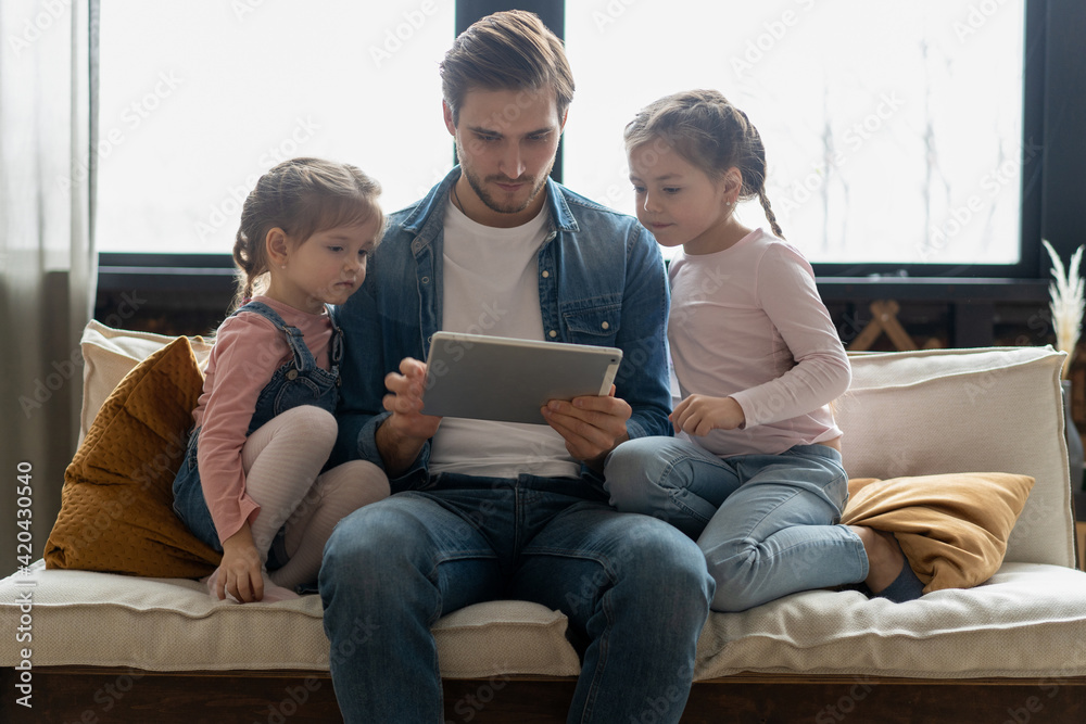 Beautiful young father, his cute little daughters are using a tablet and smiling, sitting on sofa at home.