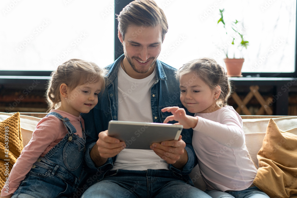 Beautiful young father, his cute little daughters are using a tablet and smiling, sitting on sofa at home. Beautiful young father, his cute little daughters are using a tablet and smiling, sitting on sofa at home.