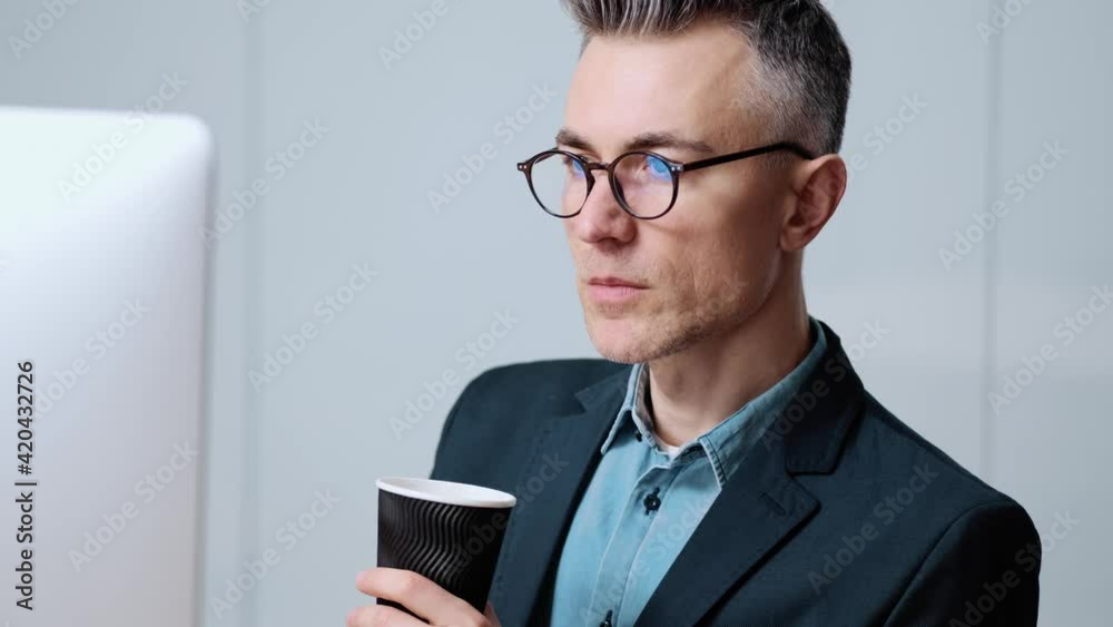 A focused businessman wearing glasses is working with his computer while drinking a cup of coffee sitting in the office