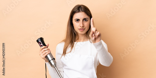Woman using hand blender over isolated background counting one with serious expression