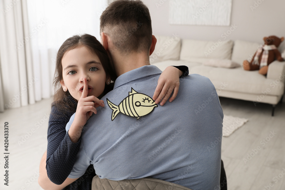 Cute little girl sticking paper fish to father's back at home - obrazy, fototapety, plakaty 