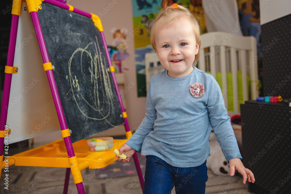 Naklejka premium A small blonde smiling girl at home in the game room near the blackboard with chalk in her hands