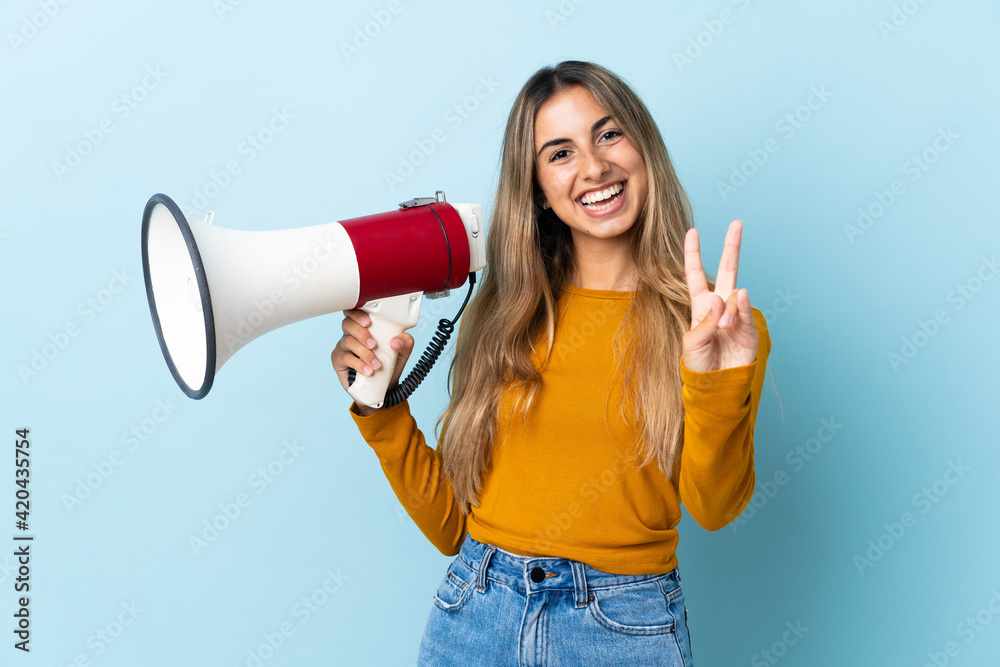 Young hispanic woman over isolated blue background holding a megaphone and smiling and showing victory sign Young hispanic woman over isolated blue background holding a megaphone and smiling and showing victory sign
