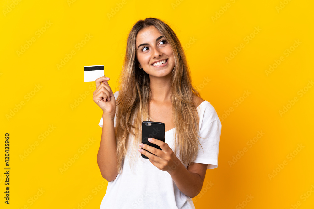 Naklejka premium Young hispanic woman over isolated yellow background buying with the mobile with a credit card while thinking