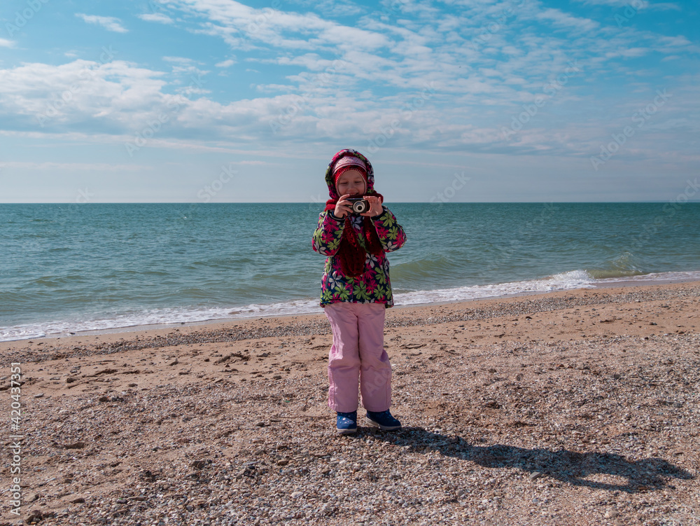 Child photographing on empty sea beach. Little girl in warm clothes taking pictures with camera in hands. Lifestyle photo Cold weather spring autumn season Family fun hobby vacation travel advertising