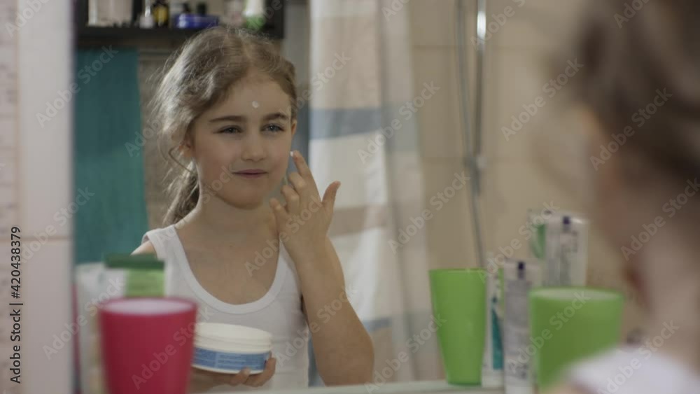 Portrait Young Funny Little Girl Applying Moisturizing Cream on Her Face in Bathroom Looking at Mirror Reflection. Happy Cute Caucasian Toothless Child Applying on Face Cream For Skin Car. Portrait Young Funny Little Girl Applying Moisturizing Cream on Her Face in Bathroom Looking at Mirror Reflection. Happy Cute Caucasian Toothless Child Applying on Face Cream For Skin Car.