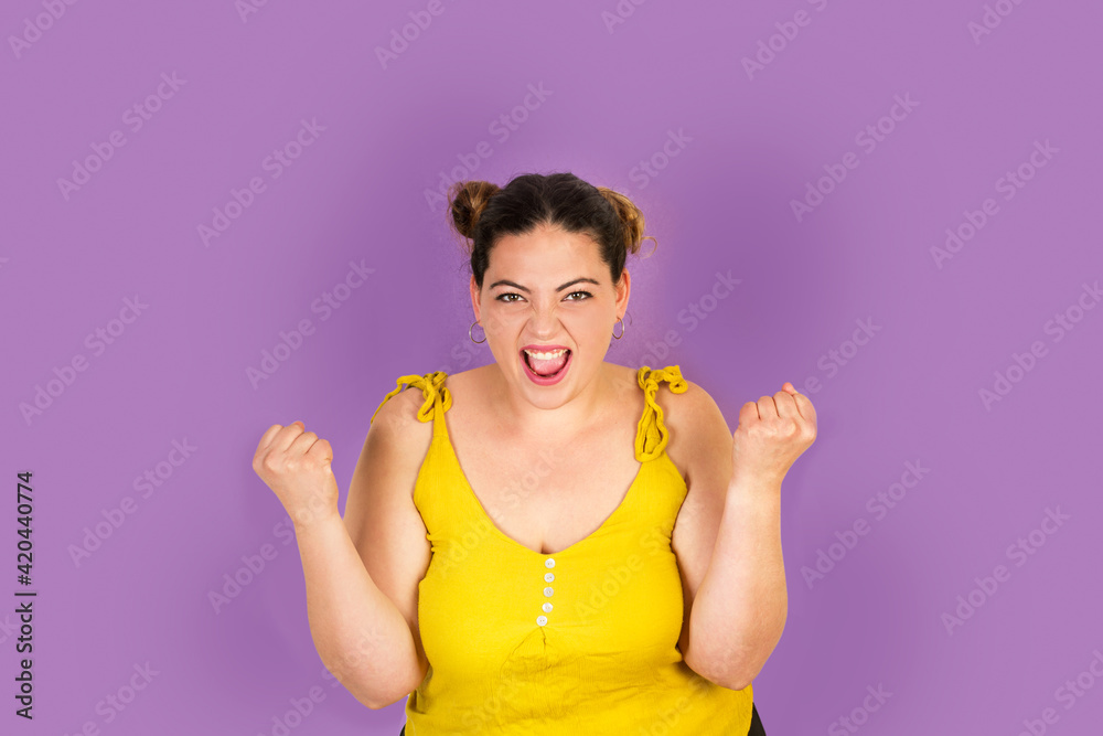 Mujer joven gritando y con las manos levantadas y los puños cerrados sobre un fondo morado liso y aislado. Vista de frente. Copy space Mujer joven gritando y con las manos levantadas y los puños cerrados sobre un fondo morado liso y aislado. Vista de frente. Copy space