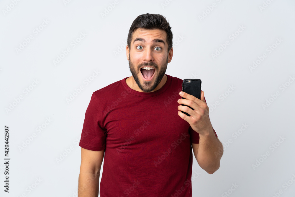 Young handsome man with beard using mobile phone isolated on white background with surprise and shocked facial expression Young handsome man with beard using mobile phone isolated on white background with surprise and shocked facial expression