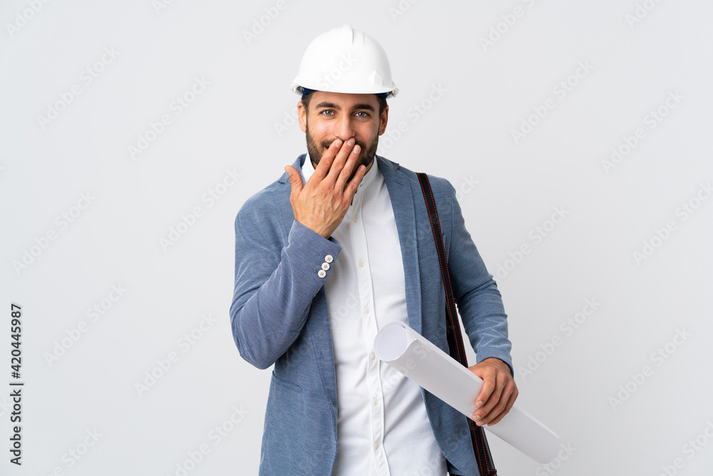 Young architect man with helmet and holding blueprints isolated on white background happy and smiling covering mouth with hand Young architect man with helmet and holding blueprints isolated on white background happy and smiling covering mouth with hand
