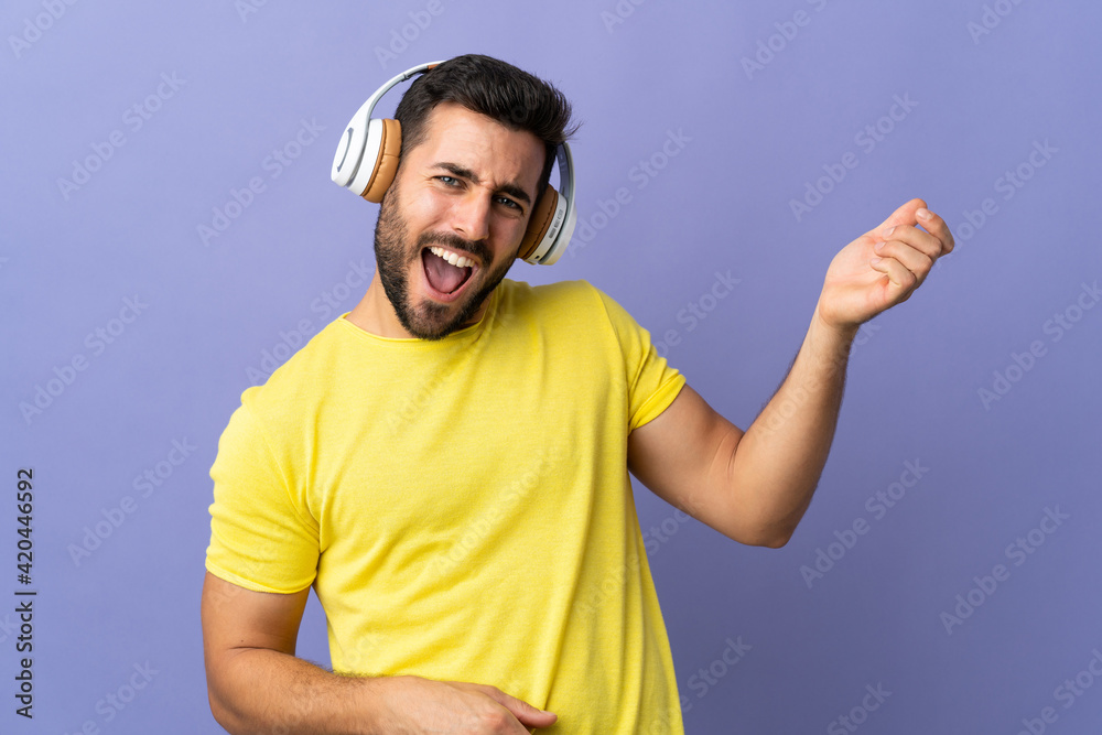 Young handsome man with beard isolated on purple background listening music and doing guitar gesture Young handsome man with beard isolated on purple background listening music and doing guitar gesture