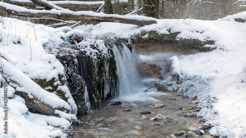 landscape with a small waterfall and flooded water, frozen icicles, stones covered with green moss, ice and snow