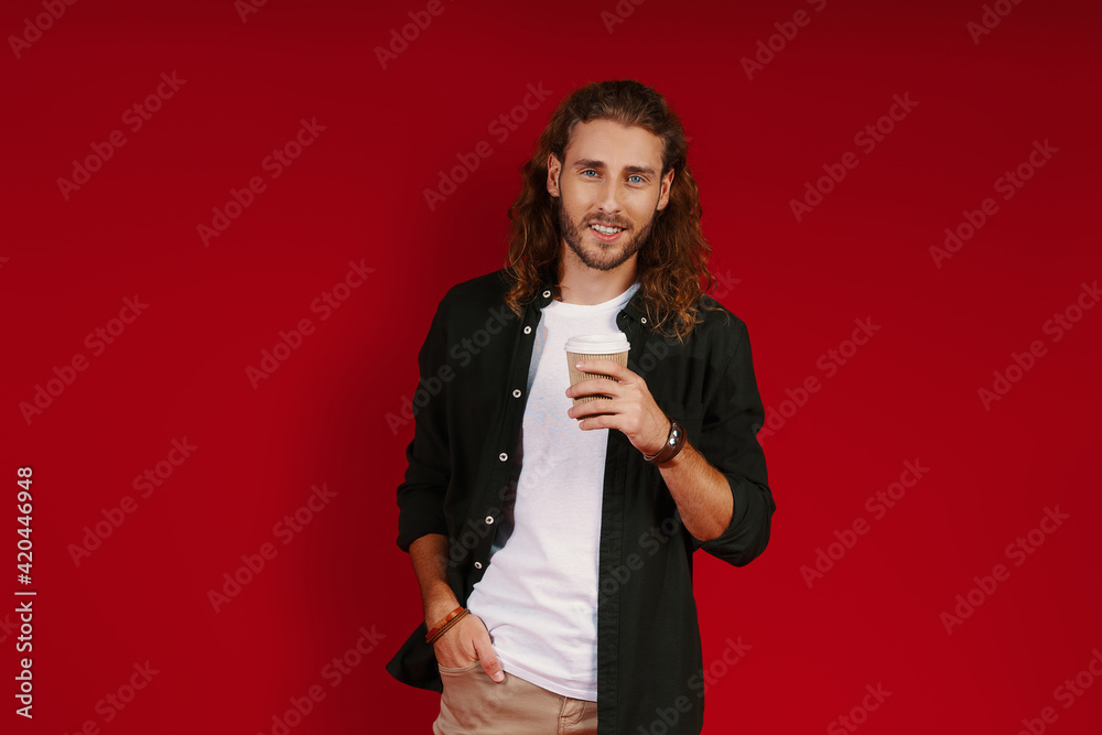 Charming young man in casual clothing looking at camera and smiling while standing against red background Charming young man in casual clothing looking at camera and smiling while standing against red background