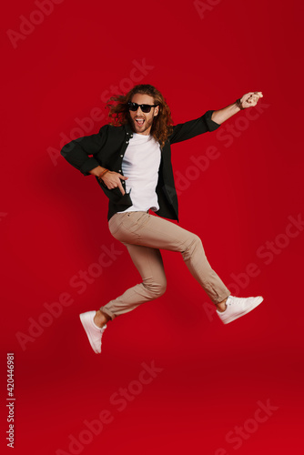 Full length of playful young man in casual clothing smiling and playing imaginary guitar while hovering against red background