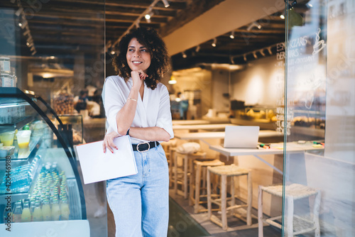 Happy female manager with menu in hand standing in doorway of local cafeteria and smiling, pretty Caucaisan small owner enjoying business lifestyle for working with franchise takeaway industry photo