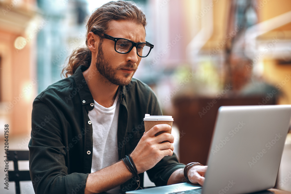 Serious young man in casual clothing working using computer and drinking coffee while sitting in cafe Serious young man in casual clothing working using computer and drinking coffee while sitting in cafe