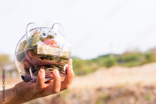 The hand holds the piggy bank, inside there are many Thai banknotes and coins.