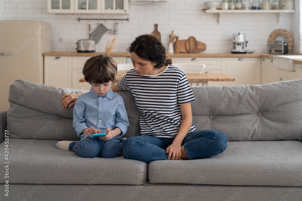 Mom relax with children on sofa enjoy video on mobile phone spending time at home together. Mother sitting on couch with little kid watch cartoons, video on smartphone. Happy loving family. 