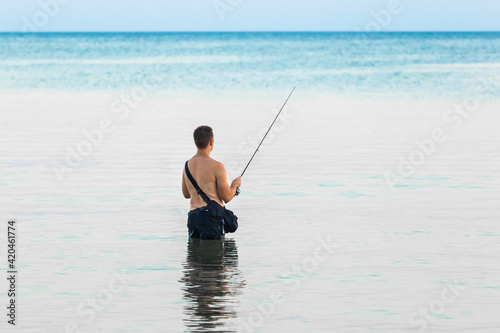 A young guy without a shirt stands knee-deep in the water of the sea in the evening and catches fish with a line