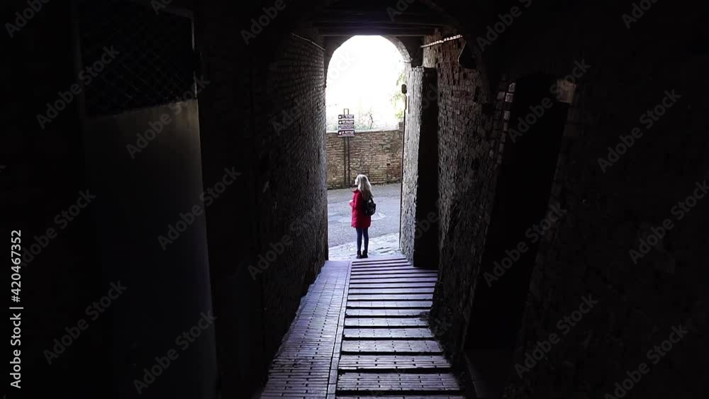 A cramped dark street in the city walls of the mid-rise city of San Gimignano. The girl in red at the end of a dark tunnel on an old street. A cramped dark street in the city walls of the mid-rise city of San Gimignano. The girl in red at the end of a dark tunnel on an old street.