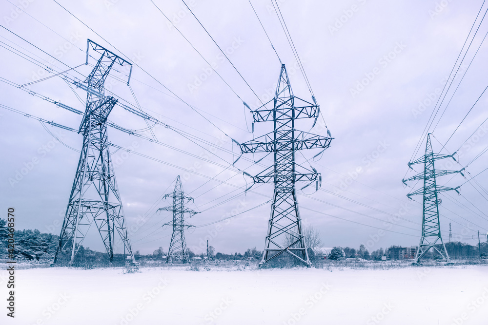 Evening view of many high-voltage towers with wires against the background of a winter snow-covered forest with elements of civilization in blue tones