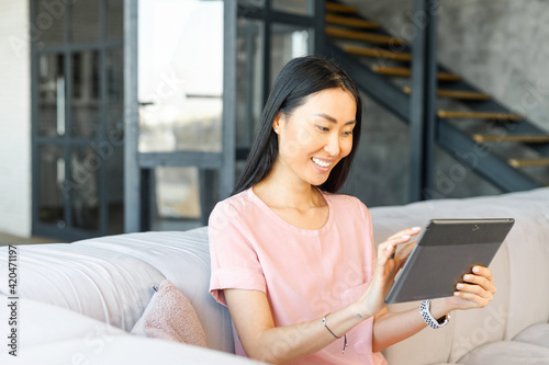 Asian woman with black straight hair using modern grey digital tablet while sitting in loft-style apartment, smiling, touching the screen, reading pleasant message from boyfriend, relaxing on sofa