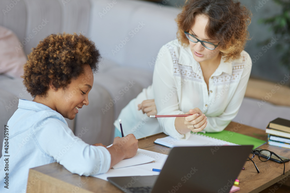 Learning excellence. Joyful mixed race teen schoolgirl writing during a lesson with her female teacher at home Learning excellence. Joyful mixed race teen schoolgirl writing during a lesson with her female teacher at home