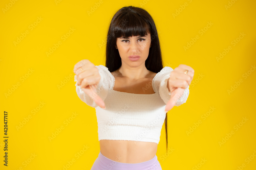 Young beautiful brunette woman wearing casual clothes standing over yellow background looking unhappy and angry showing rejection and negative with thumbs down gesture. Bad expression. Young beautiful brunette woman wearing casual clothes standing over yellow background looking unhappy and angry showing rejection and negative with thumbs down gesture. Bad expression.