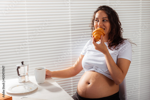 Happy pregnant young beautiful woman eating croissant and looking througt blinds during morning breakfast. Concept of pleasant morning and positive attitude during pregnancy photo