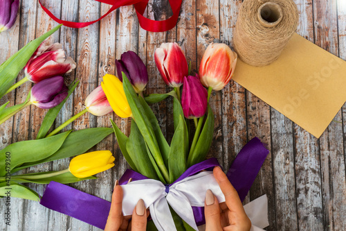 Florist decorating bouquet, close-up. Hands tying ribbon on bouquet of flowers indoors on wooden background, selective focus. Small business. Woman florist making a bouquet of fresh colorful tulips