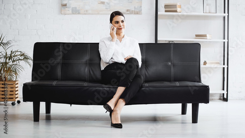 elegant businesswoman sitting on black sofa and talking on mobile phone