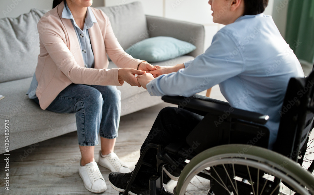 Cropped view of disabled tenager in wheelchair holding hands with female psychologist, closeup
