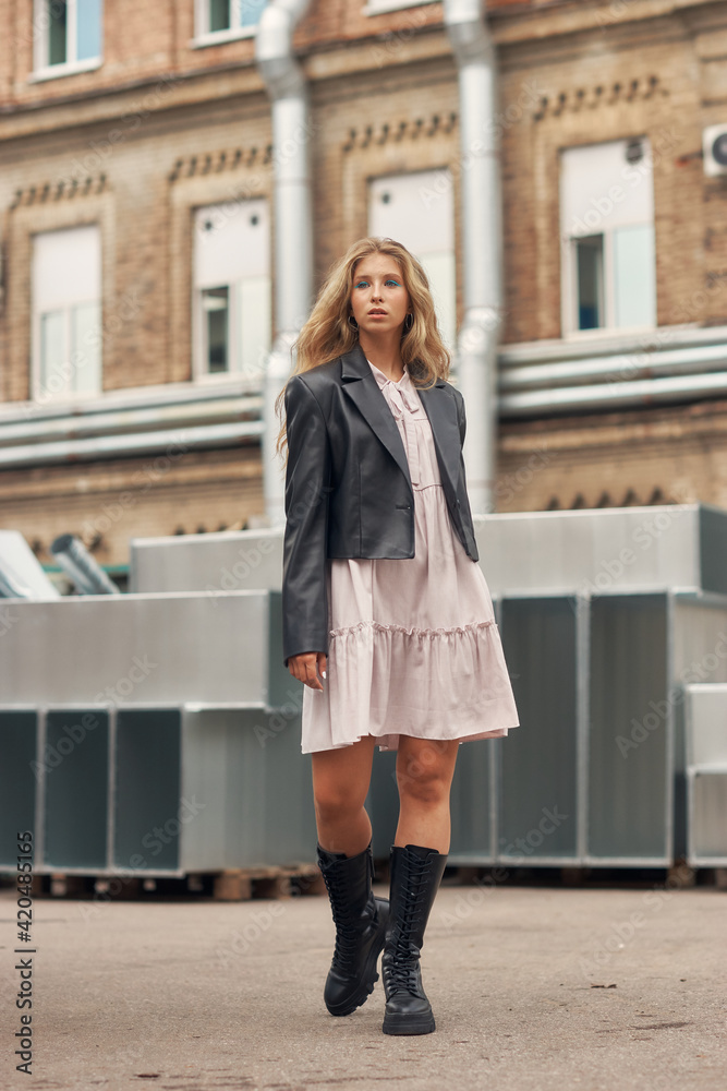 Young stylish woman in pink dress, black leather jacket and boots walking at urban location. Female model with long wavy blonde hair outdoor fashion look. Young stylish woman in pink dress, black leather jacket and boots walking at urban location. Female model with long wavy blonde hair outdoor fashion look.