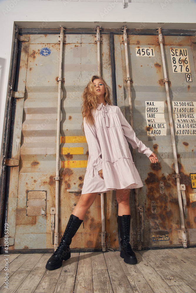 Foto Stock Young fashionable girl in pink dress and high black leather boots  standing and posing against grey metal gates. Full length. Hard light  portrait. Female model with wavy blonde hair and