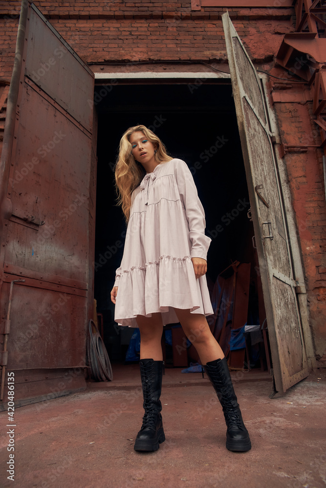 Young beautiful stylish caucasian teenager woman in pink dress and black boots posing outdoors against old factory gates. Urban portrait