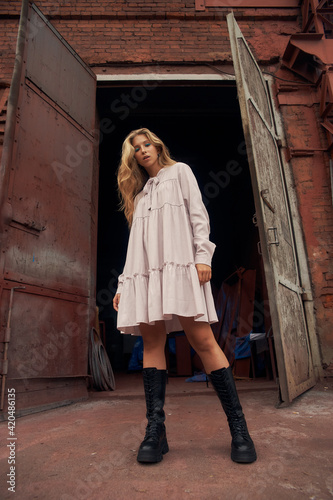 Young beautiful stylish caucasian teenager woman in pink dress and black boots posing outdoors against old factory gates. Urban portrait