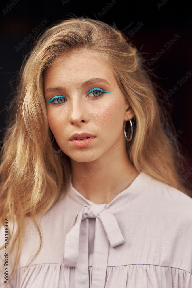 Close-up outdoor portrait. Stylish teenaager girl with long blonde hair and blue eye arrows in pink dress posing outdoors against black background Close-up outdoor portrait. Stylish teenaager girl with long blonde hair and blue eye arrows in pink dress posing outdoors against black background
