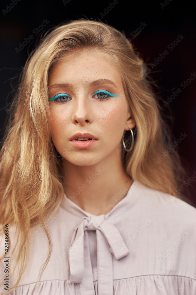 Close-up outdoor portrait. Stylish teenaager girl with long blonde hair and blue eye arrows in pink dress posing outdoors against black background Close-up outdoor portrait. Stylish teenaager girl with long blonde hair and blue eye arrows in pink dress posing outdoors against black background