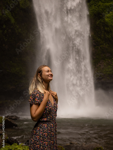 Happy woman smiling and enjoying waterfall landscape. Nature and environment concept. Travel lifestyle. Woman wearing dress. Copy space. Nung Nung waterfall in Bali