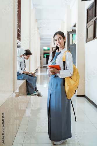 full body shot of a high school girl smiles at the camera holding a note book and wearing a school bag in the background of a building corridor.