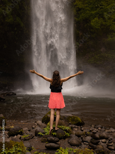 Traveler woman wearing pink dress at waterfall. Excited woman raising arms in front of waterfall. Travel lifestyle. Energy of water. View from back. Copy space. Nung Nung waterfall in Bali