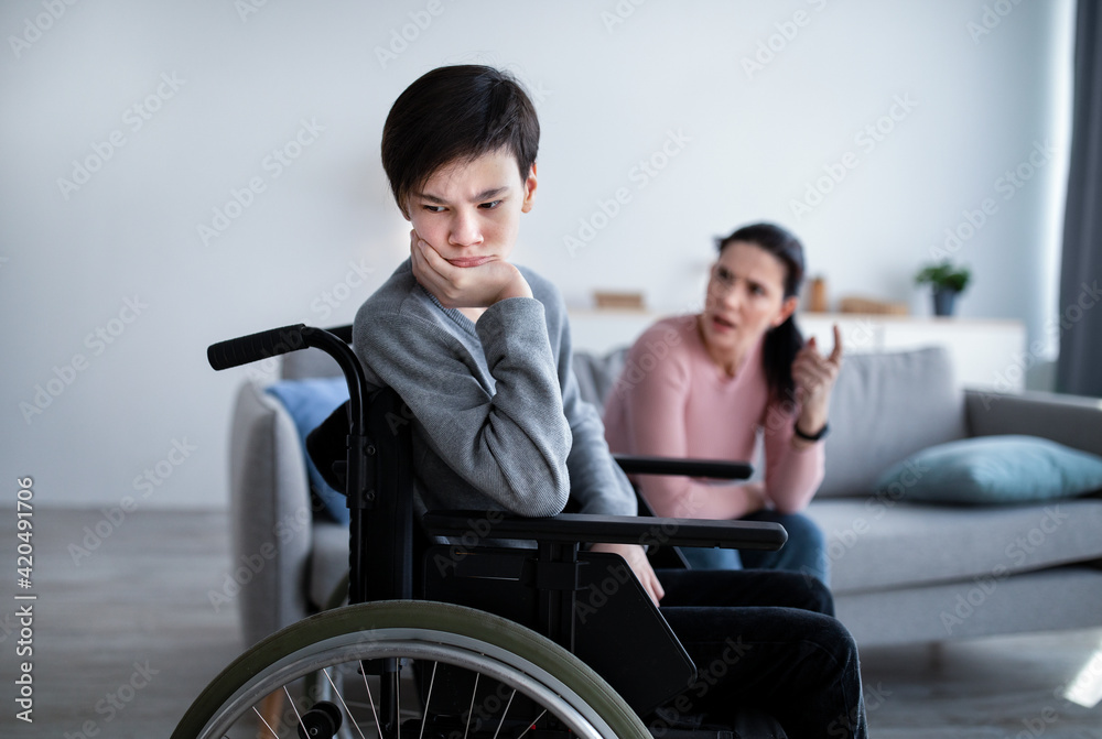 Unhappy teen boy in wheelchair having conflict with his mother, listening to her scolding at home. Relationship problems Unhappy teen boy in wheelchair having conflict with his mother, listening to her scolding at home. Relationship problems