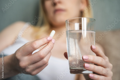 Stressed woman drinking white round pill while sitting in bed with glass of water in hand.