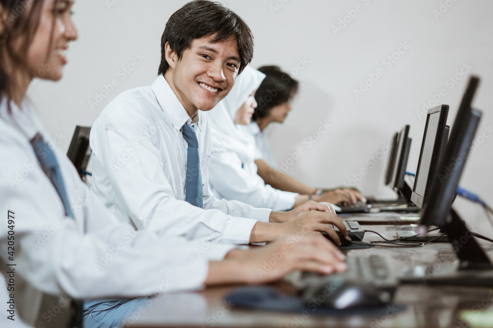 Obraz premium Male high school students smile looking at the camera while using a computer PC with their friends studying in a computer practice room at school