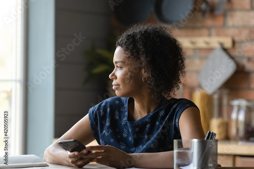 Pensive millennial biracial female take pause in paperwork ponder on offer received in phone message. Thoughtful young black lady look at window hold cell plan business conversation before making call