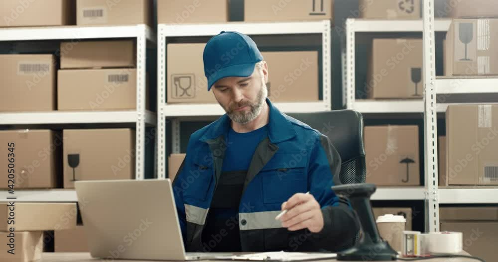 Senior warehouse worker in uniform sitting at desk folds cardboard boxes, preparing parcels for departure making notes on paper. Senior warehouse worker in uniform sitting at desk folds cardboard boxes, preparing parcels for departure making notes on paper.