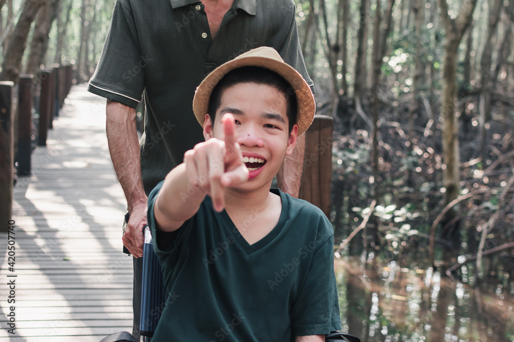 Fototapeta premium Asian special child on wheelchair pointing finger forward on mangrove forest background,Activity outdoor with family on travel, Lifestyle in the education age of disabled children,Happy disabled kid.